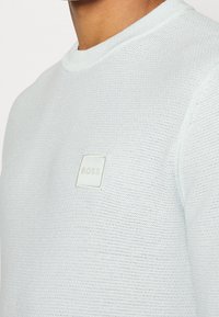 Hellblauer Strickpullover mit texturiertem Finish, rundem Halsausschnitt und kleinem quadratischen Logo-Patch mit der Aufschrift "BOSS" auf der Brust.