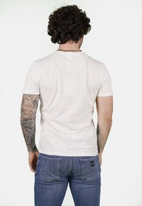 T-shirt bianco a maniche corte con scollo rotondo, indossato con jeans blu. Tessuto leggermente strutturato; tatuaggi visibili sulle braccia.
