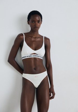 Witte lingerie set met een bralette met een zwart, gemarkeerd elastieken band en bijpassende hoge slip met roze borduursel. Glad materiaal.