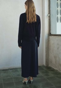 Námornícky pletený sveter v kombinácii s dlhou, jednofarebnou námorníckou sukňou a bielymi sandálmi na podpätku, zobrazený zozadu na neutrálnom pozadí.