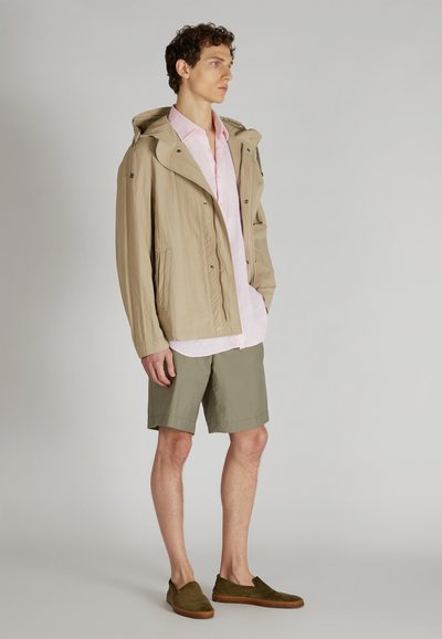 Beżowa, lekka kurtka z kapturem, jasnoróżowa koszula zapinana na guziki, oliwkowe szorty i ciemnozielone buty wsuwane. Gładka tekstura, casualowy design.