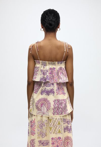 Femme aux cheveux foncés coiffés en chignon, portant une robe crème avec des motifs floraux et cachemire roses et violets, vue de dos sur fond blanc.