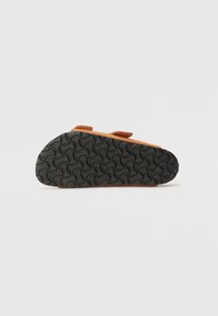 Chaussure en daim marron avec deux bandes Velcro ajustables. La semelle extérieure en caoutchouc noir présente un motif de semelle ondulé pour une bonne adhérence et traction.