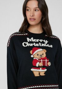 Schwarzer Strickpullover mit einem zentralen Weihnachtsteddybären, der ein Geschenk hält. Darüber steht der Text "Frohe Weihnachten" mit roten und weißen Mustern an den Ärmeln.