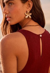 Bordeauxrode tanktop met een sleutelgatdetail aan de achterkant. Het model draagt een grote gouden bloemige oorbel en een korte kralenhalsketting. Gladde textuur.