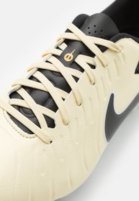 Tej színű sportcipő, texturált felülettel, fekete részletekkel és fehér Nike swoosh-szal. Lapos fűzőkkel és perforációkkal rendelkezik a nyelvén.