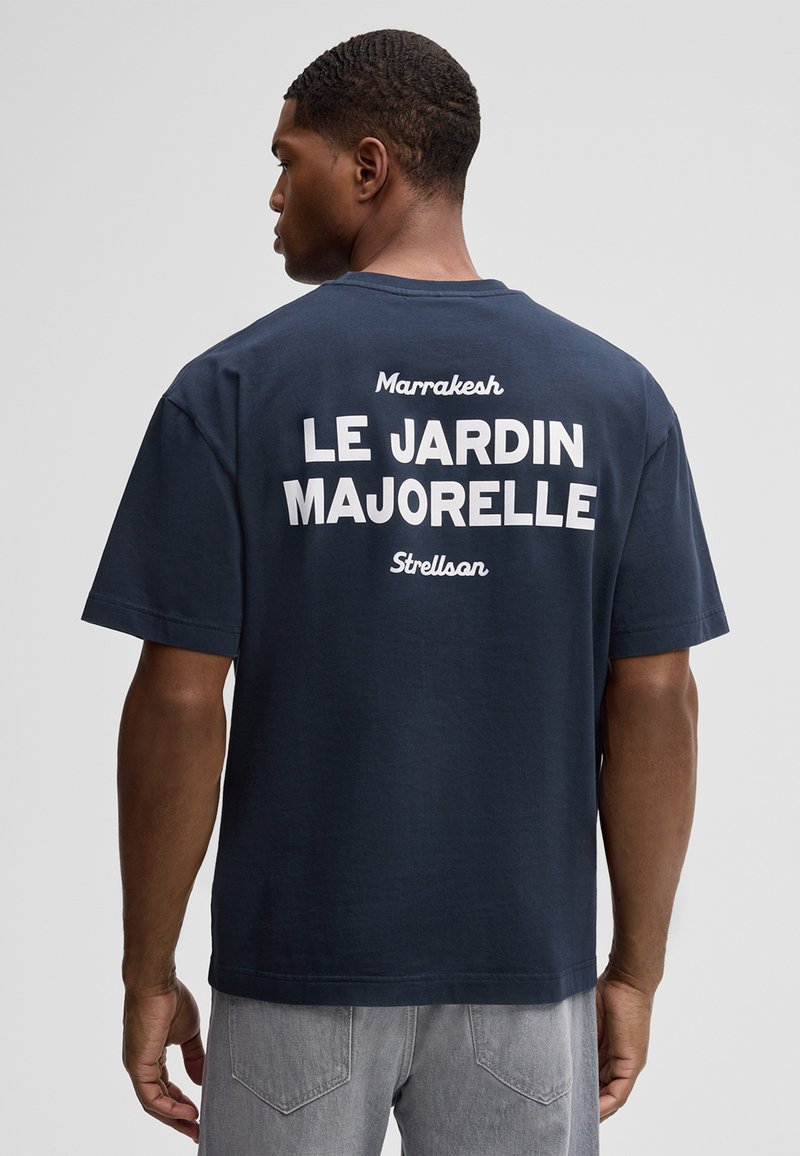 Man draagt een marineblauw T-shirt met witte tekst "Marrakesh LE JARDIN MAJORELLE Strellson" op de achterkant, gecombineerd met grijze spijkerbroek.