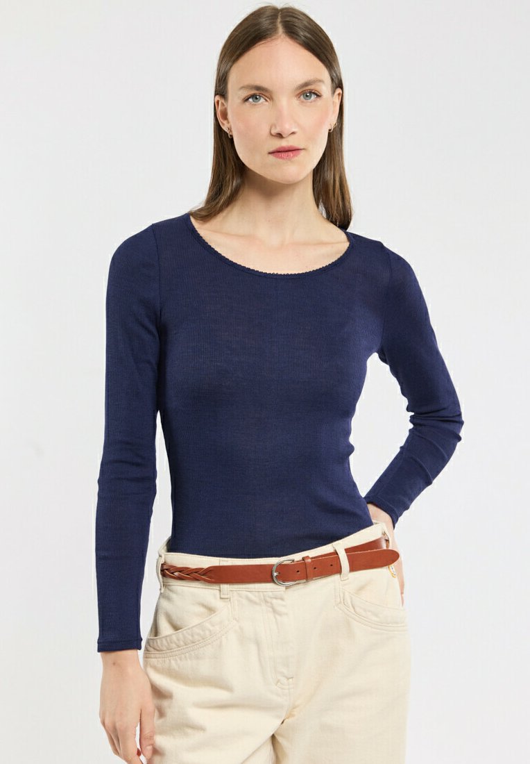 Femme aux cheveux bruns lisses portant un haut à manches longues bleu marine et un pantalon beige taille haute avec une ceinture tressée marron, se tenant devant un fond blanc.