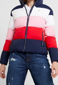 Puffer jacket with a hood featuring horizontal stripes in navy, white, pink, and red. Two zippered side pockets and a front zipper.