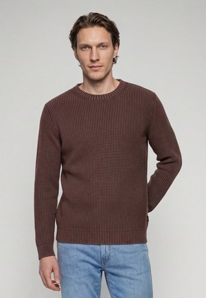 Mand iført en brun ribstrikket sweater og lyseblå jeans, stående mod en ensfarvet lys baggrund med den ene hånd bag ryggen.