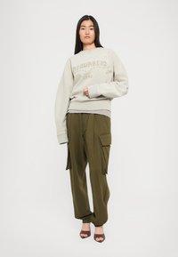 Woman wearing beige DSQUARED2 sweatshirt and olive cargo pants with brown open-toe heels, standing against a white background.