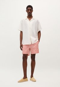 Chemise blanche à manches courtes avec un tissu texturé, boutons beiges, assortie à un short à motifs rouges et des espadrilles beiges.