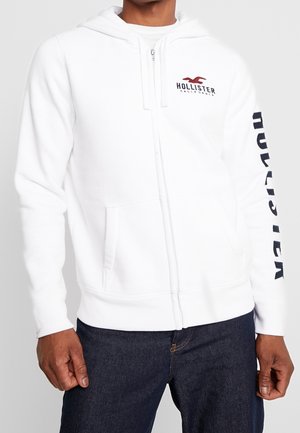 Homme portant un sweat à capuche blanc zippé avec le logo Hollister sur la poitrine et la manche, associé à un jean bleu foncé.