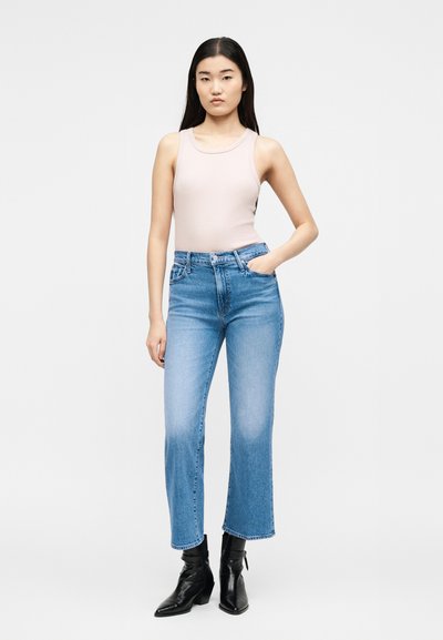 Γυναίκα στέκεται με το ένα χέρι στην τσέπη, φοράει ανοιχτό ροζ αμάνικη μπλούζα, μπλε ψηλόμεσο τζιν και μαύρα μποτάκια σε λευκό φόντο.