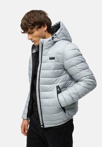 Lys grå pufferjakke med horisontal quiltet design, fast hætte og sorte lynlåse. Har sidelommer og diskret branding.