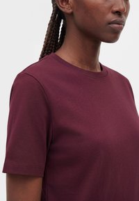 Femme aux cheveux foncés tressés portant un t-shirt uni bordeaux à manches courtes et col rond, montrée des épaules au menton sur un fond gris.