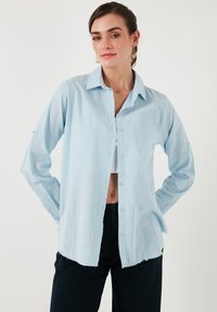 Camicia azzurro chiaro a maniche lunghe con bottoni, dotata di taschino, con una texture sottile e orlo sfilacciato. Abbinata a pantaloni neri.