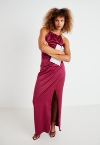 Μπορντό σατέν φόρεμα με λαιμόκοψη halter και πλευρική σχισμή, λεία υφή και εφαρμοστή σχεδίαση. Συνοδευόμενο από ασημένια τσάντα και πέδιλα με λουράκια.