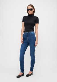 Black ribbed turtleneck top paired with dark blue skinny jeans. The model wears large black sunglasses and black flat shoes.