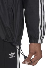Černá větrovka s bílými pruhy na rukávech, elastickými manžetami, nastavitelným spodním lemem se stahovací šňůrkou, doplněná černými kalhotami s logem Adidas.
