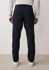 Námornícke nohavice vyrobené z textúrovanej tkaniny, s dvoma zadnými vreckami, strihom na mieru a v kombinácii s bielymi teniskami na neutrálnom pozadí.