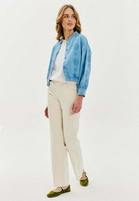 Světle modrá vyšívaná bunda, bílá košile, krémové kalhoty a zelené boty s bílými akcenty. Model stojí na pozadí bez vzoru.