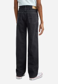 Crne široke traperice s visokim strukom, koje imaju dva strašnja džepa i svjetlosmeđi natpis na stražnjem dijelu. Teksturirani traper s laganim šavovima.