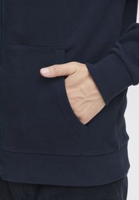 Marineblå zip hoodie med ribbet mansjetter og kant, med en hånd i frontlommen, laget av myk teksturert stoff.