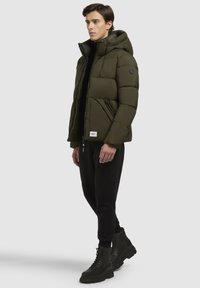 Chaqueta puffer verde olivo con capucha, que cuenta con bolsillos con cremallera y un parche con el logo. Lleva puesta sobre un cuello de tortuga negro y pantalones negros con botas gruesas.