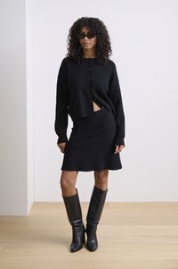 Čierny kratší cardigan s gombíkmi, kombinovaný s čiernou priliehavou sukňou. Modelka má na sebe čierne kožné čižmy po kolená a slnečné okuliare.