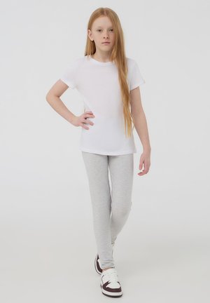 Biała bawełniana koszulka, szare legginsy i białe sneakersy z burgundowymi akcentami. Modelka pozuje z ręką na biodrze, ma długie proste włosy.