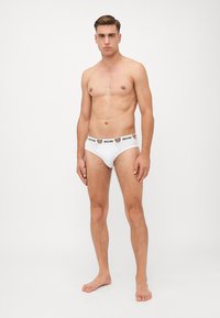 Weiße Baumwollunterhosen mit einem schwarzen Bund, der das Moschino-Logo und eine Bärengrafik zeigt. Das Modell steht barfuß in einer neutralen Pose.
