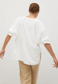 Camicia bianca ampia con maniche arrotolate, scollatura rotonda e spacchi laterali. Tessuto morbido con una superficie liscia, abbinata a pantaloni beige.