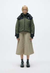 Casaco acolchoado verde com detalhes pretos, saia bege acolchoada e sapatos de caminhada pretos, contra um fundo simples.