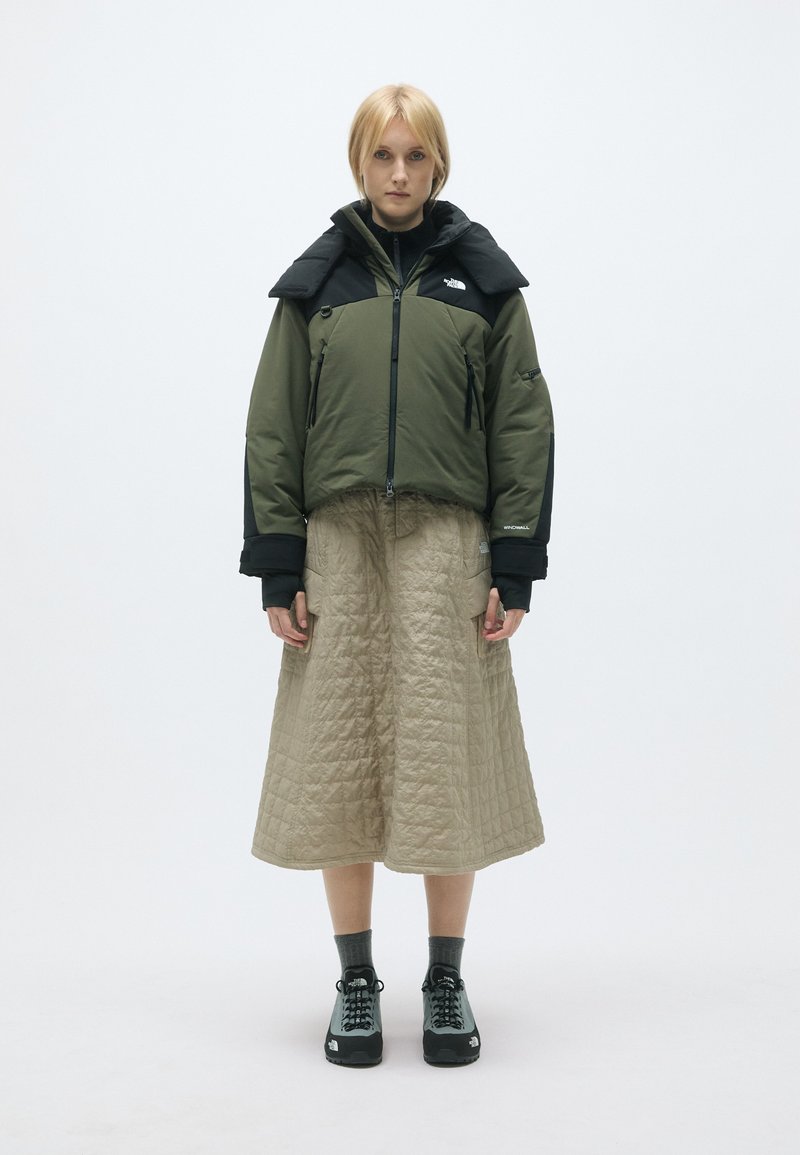 Casaco acolchoado verde com detalhes pretos, saia bege acolchoada e sapatos de caminhada pretos, contra um fundo simples.