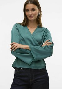 Teal indpakningsbluse med lange klokkeslætærmer, med blonder trim langs halsudskæringen og ærmerne, parret med mørkeblå jeans.