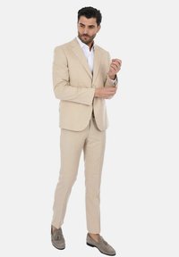 Completo beige con un motivo testurizzato, design slim fit, giacca monopetto e pantaloni abbinati. Accompagnato da una camicia bianca e scarpe grigie.