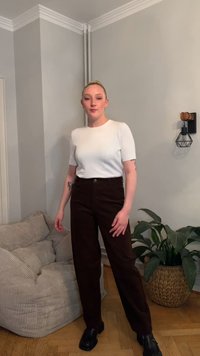 Bílý top s krátkým rukávem, hnědé manšestrové kalhoty a černé boty. Stojí opřený o světle šedou zeď, poblíž je vzorované polstrované křeslo a rostlina.