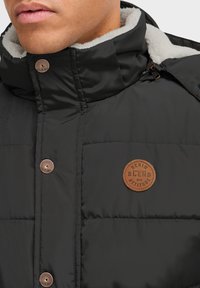 Černá prošívaná bunda s plyšovým bílým límcem. Obsahuje bronzové kování a kulatou koženou nášivku s textem "Denim Blend with Attitude".