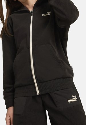 Femme portant une veste polaire noire zippée Puma et un pantalon assorti avec des logos Puma blancs sur la poitrine et la cuisse, sur un fond neutre.