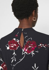 Blusa preta com padrão floral, apresentando rosas vermelhas e brancas, detalhe de decote em forma de chave nas costas e fechamento com botão no decote, confeccionada em tecido macio.