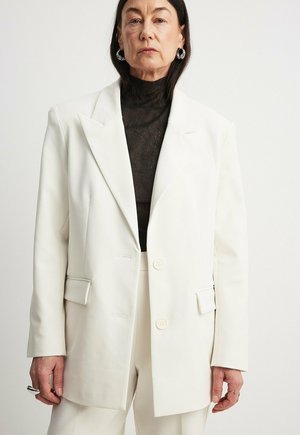 Blazer blanc surdimensionné avec col en pointe, deux boutons et deux poches frontales, superposé sur un haut texturé noir. Design minimaliste.