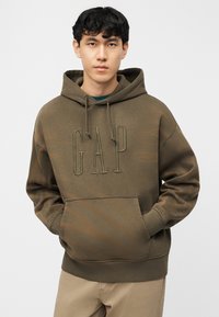 Νέος άνδρας που φοράει πράσινο χακί φούτερ GAP με τις χέρια στις μπροστινές τσέπες, στέκεται μπροστά σε ένα απλό φωτεινό φόντο.