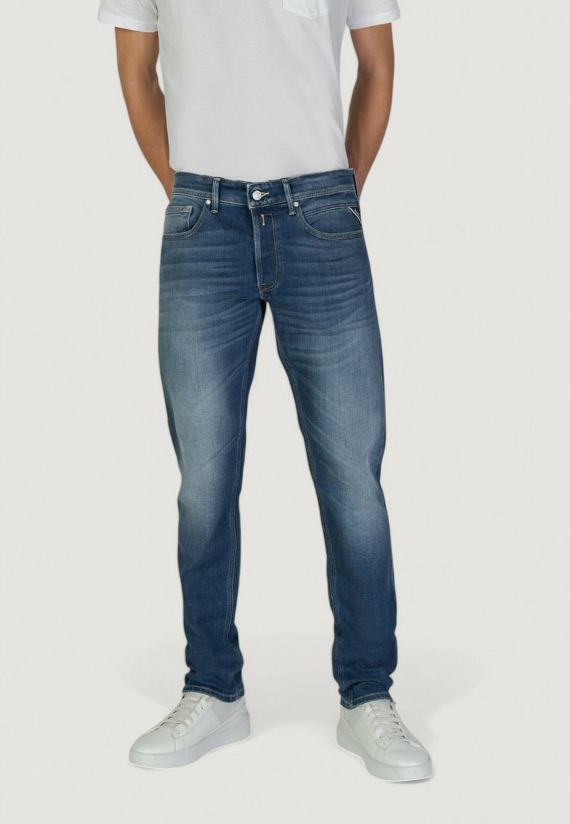Jeans in denim blu scuro con taglio dritto, leggero sbiadimento sulle cosce e design a cinque tasche, abbinati a sneakers bianche e a una maglietta grigia.