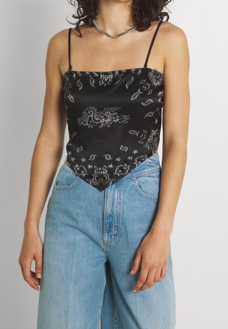 Haut en satin noir avec motifs blancs complexes de dragon et cachemire, doté de bretelles fines et d'un ourlet pointu, associé à un jean bleu clair.