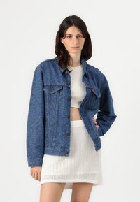 Denim jacket with intricate blue paisley pattern, oversized fit, front buttons, chest pockets, worn over a light-colored crop top and textured skirt.