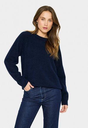 Femme aux longs cheveux bruns portant un pull bleu marine et un jeans bleu foncé, posant avec une main dans la poche sur fond blanc.