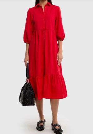 Nainen pukeutuneena punaiseen napitettuun midipituiseen mekkoon, jossa on puffyhihat, pitää käsissään mustaa niitillä koristeltua käsilaukkua ja käyttää mustia solkikenkiä.