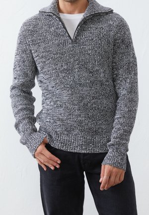 Muž oblečený v šedém a černém pleteném svetr s čtvrtinovým zipem přes bílou košili, v kombinaci s černými kalhotami, ruce volně podél těla.