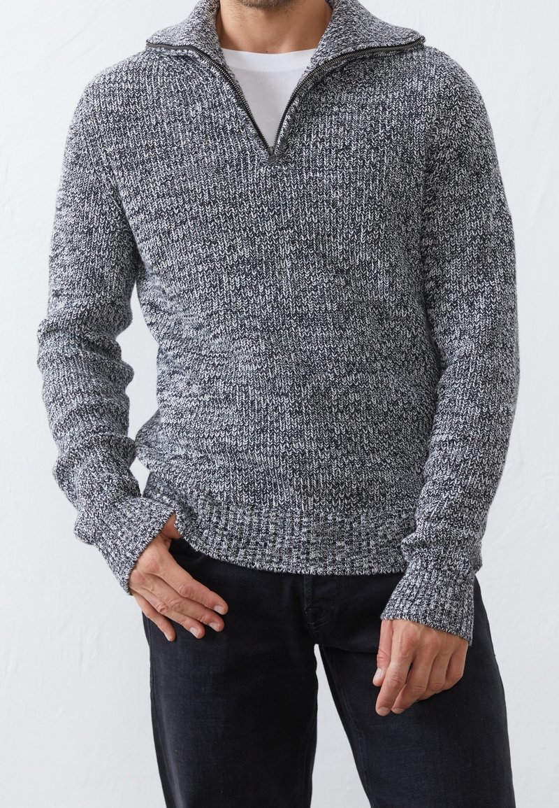 Homme portant un pull en maille gris et noir à fermeture éclair quart de zip sur une chemise blanche, associé à un pantalon noir, les mains relâchées le long du corps.